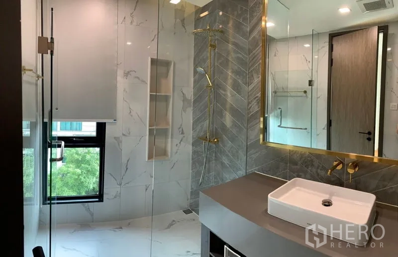 Condominium for sale in Khlong Toei, Bangkok - Modern bathroom with marble tiles, gold fixtures, glass shower enclosure and a square basin vanity.
