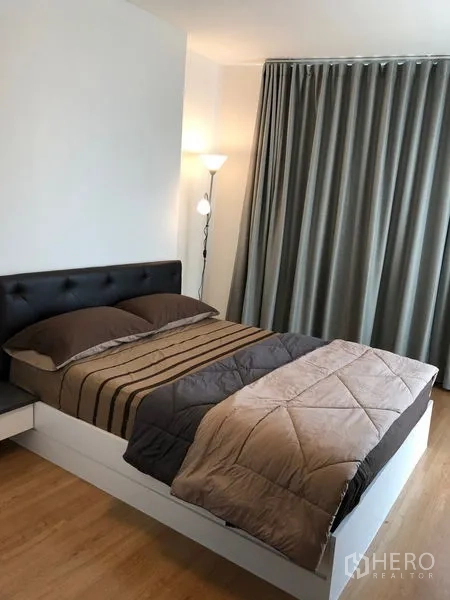Condominium for rent in Huai Khwang, Bangkok - Bedroom with double bed, floor lamp, and full-length blackout curtains on wood floors.