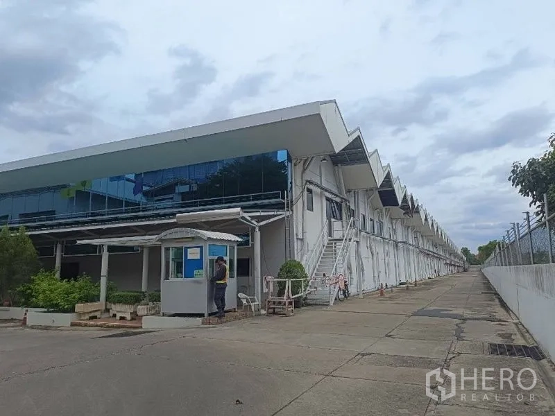 货栈 出租在Lam Luk Ka, Pathum Thani - 仓库前端玻璃办公区与门卫亭，侧面服务楼梯沿建筑延伸。
