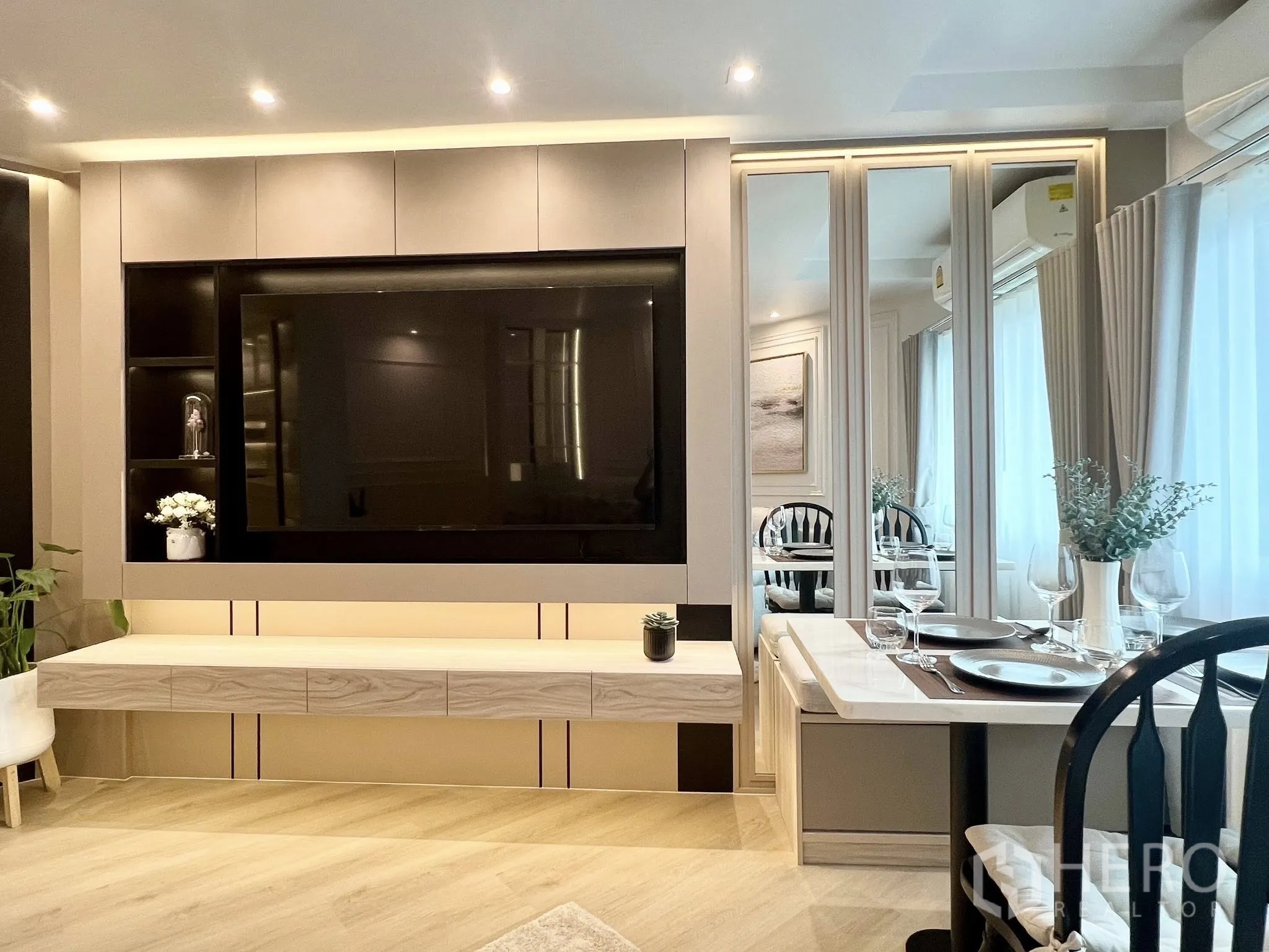 Condominium for rent or sale in Yan Nawa, Bangkok - Living area with a large built-in TV feature wall, mirrored panels and a set dining table by the window.