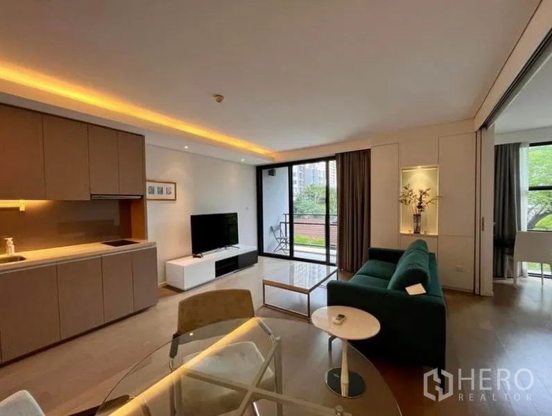 Condominium for rent in Watthana, Bangkok - Bright living room with open kitchen, green sofa, TV console, and a balcony overlooking leafy views.