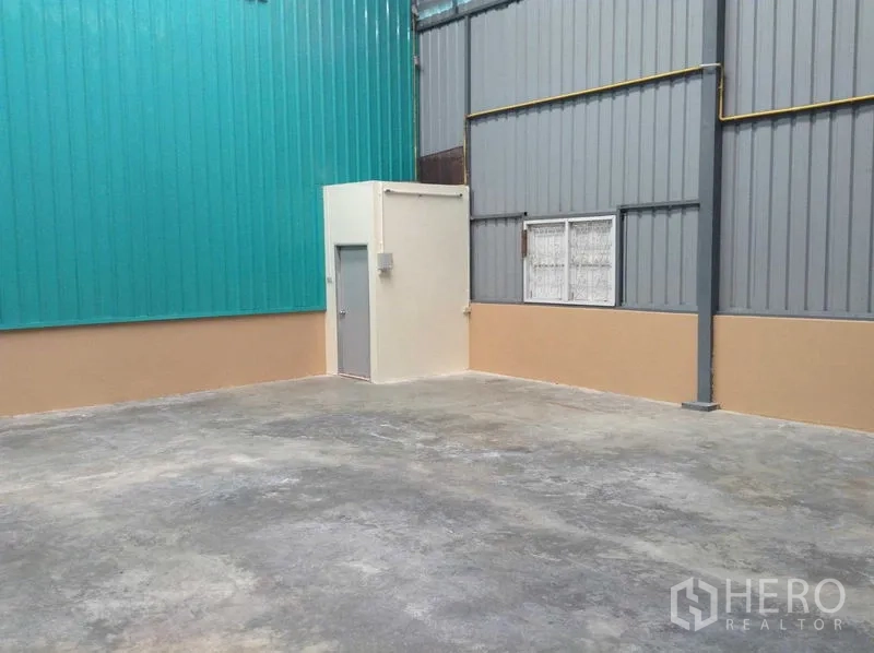 Warehouse for rent in Bang Phli, Samut Prakan - Concrete yard beside teal and gray metal-clad warehouse walls with a small utility room.
