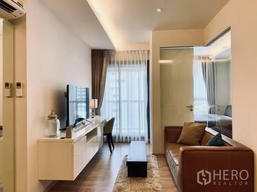 Condominium for rent in Watthana, Bangkok - Bright living room with leather sofa, TV console, and floor‑to‑ceiling window, with glass partition to the bedroom.