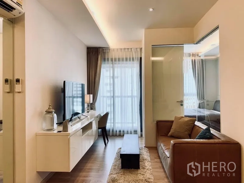 Condominium for rent in Watthana, Bangkok - Bright living room with leather sofa, TV console, and floor‑to‑ceiling window, with glass partition to the bedroom.