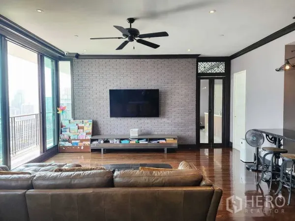 Condominium for sale in Khlong Toei, Bangkok - Living area with brick feature wall, wall-mounted TV and ceiling fan opening to a balcony.