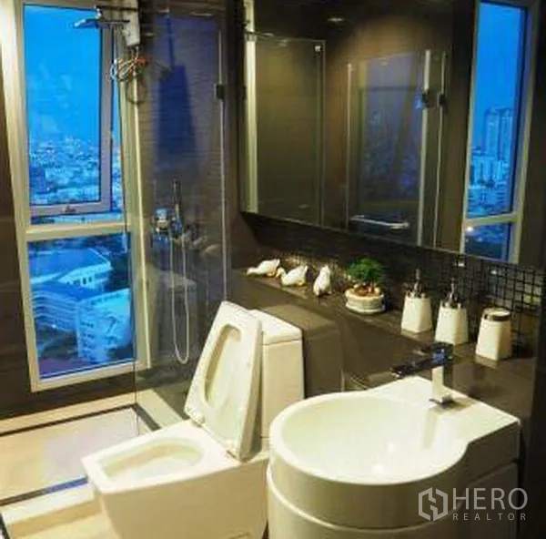 Condominium for rent in Sathon, Bangkok - Modern bathroom with glass shower, large window and vanity with city view.