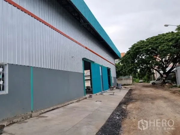 货栈 出租在Saphan Sung, Bangkok - 仓库侧立面，多个装卸门与混凝土车道，临绿树成荫的小路。