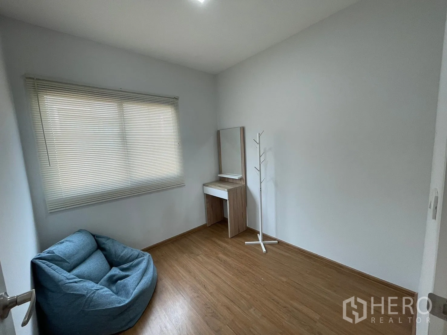 Townhouse for rent in Bang Phli, Samut Prakan - Bright spare bedroom with large window, white blinds, wood flooring, a blue bean bag chair, and a dressing table.