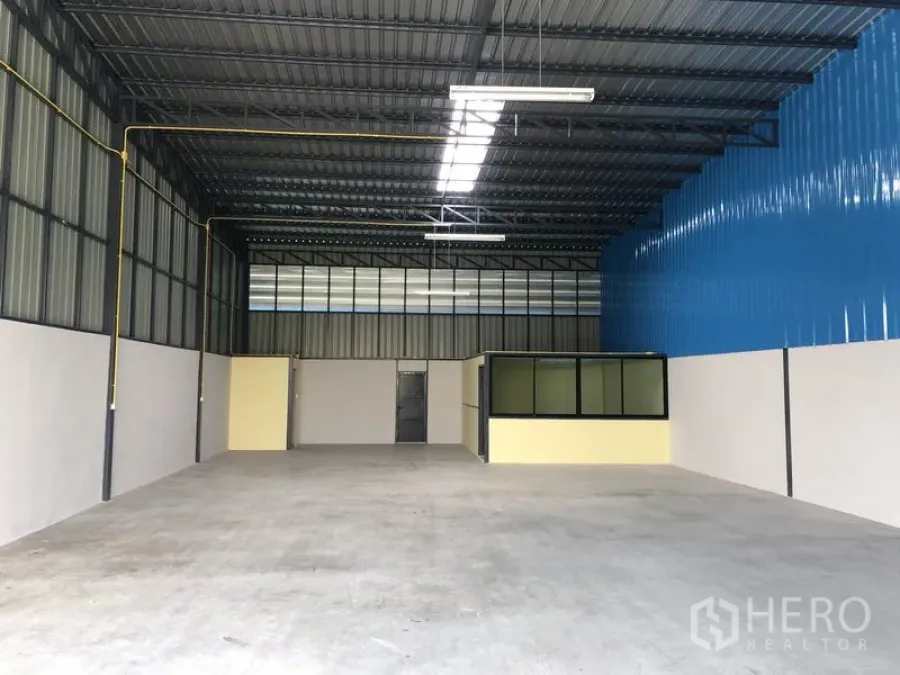 Warehouse for rent in Khlong Luang, Pathum Thani - Interior of a clean warehouse showing high steel truss roof with skylights, smooth concrete floor and a small glass-partitioned office.
