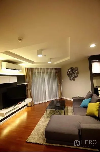 Condominium for rent in Huai Khwang, Bangkok - Living area with sectional sofa, rug, glass coffee table and floor-to-ceiling curtains.