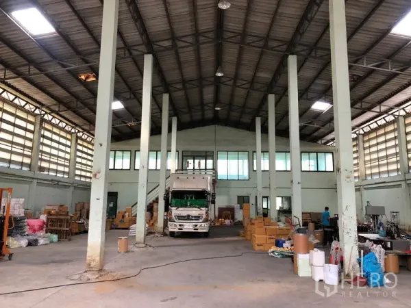 货栈 出租在Nonthaburi - 仓库内部高层空间，钢梁屋顶与立柱，货车停靠，地面堆放纸箱与货架。