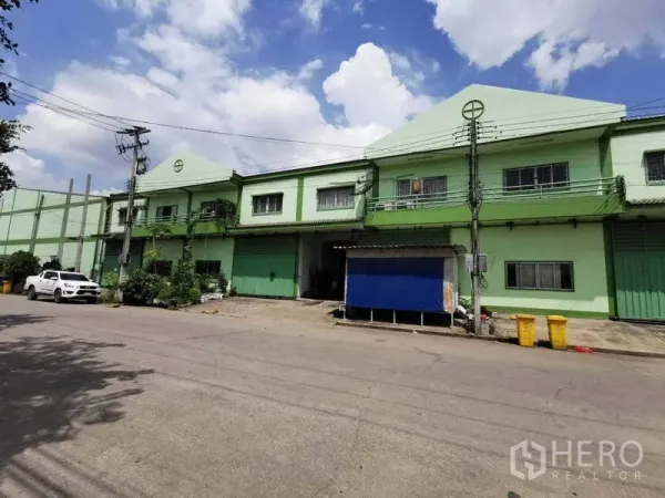 โรงงาน ให้เช่าในSamut Sakhon - มุมถนนโชว์อาคารโรงงานสองชั้นสีเขียวอ่อน ประตูม้วน ระเบียง และพื้นที่จอดรถ