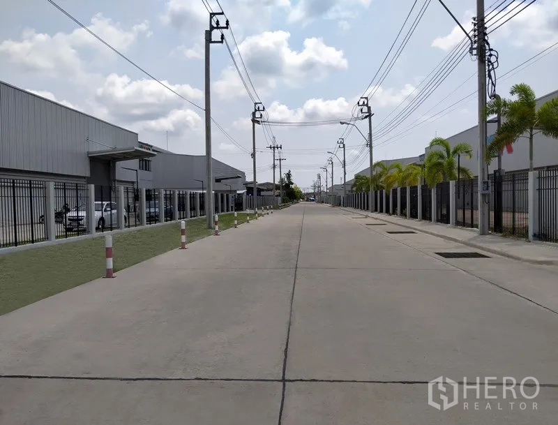 Factory for rent or sale in Samut Prakan - Wide internal road of the industrial estate with power lines and gated factories.
