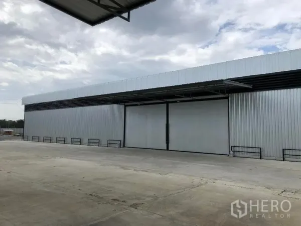 Warehouse for rent in Pathum Thani - Front exterior of a large metal-clad warehouse with wide sliding doors and a spacious concrete loading yard under cloudy skies.