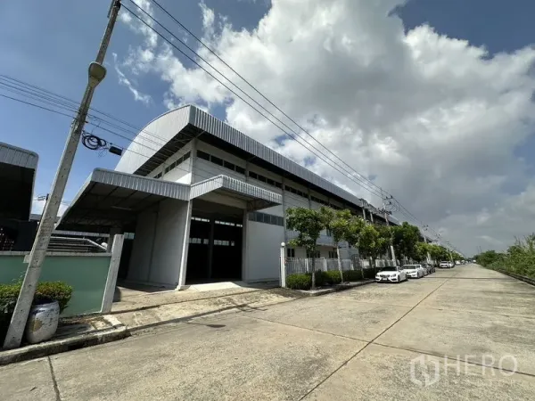 工厂 出租在Lam Luk Ka, Pathum Thani - 金属板外墙的大型工厂侧视图，街道两旁有树与停车位。