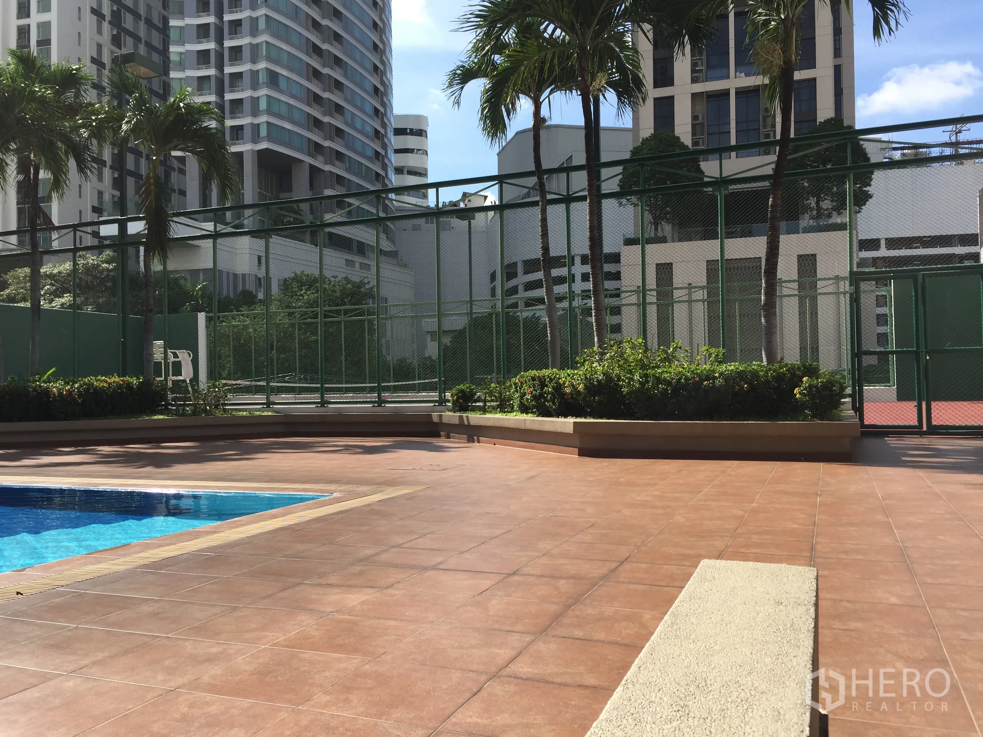 Condominium for rent in Watthana, Bangkok - Outdoor swimming pool deck beside fenced tennis courts and palm trees.