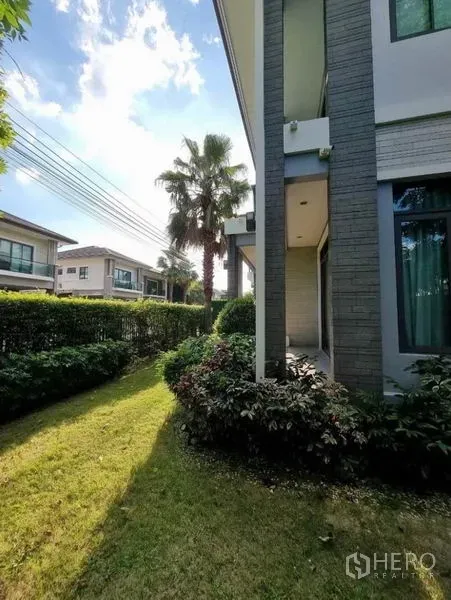 Detached House for sale in Suan Luang, Bangkok - Corner garden along the side of the home with low hedges, a palm tree and neighboring houses.