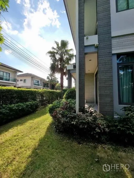 Detached House for sale in Suan Luang, Bangkok - Corner garden along the side of the home with low hedges, a palm tree and neighboring houses.