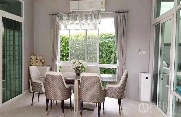 Detached House for rent in Don Mueang, Bangkok - Bright dining area with a six-seat table, chandelier and a large window facing green hedges.