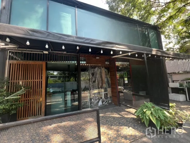 零售空间 出租在Watthana, Bangkok - 双层玻璃立面的零售店面，带遮篷灯串、露台休息区与绿植入口。