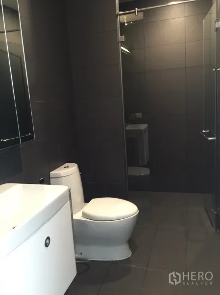 Condominium for sale in Pathum Wan, Bangkok - Second bathroom with large mirror, white vanity, and glass-enclosed shower against charcoal tiles.