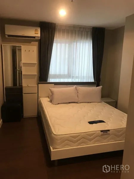 Condominium for rent or sale in Huai Khwang, Bangkok - Master bedroom with queen bed, air conditioner, wardrobe and soft lighting by a large window.