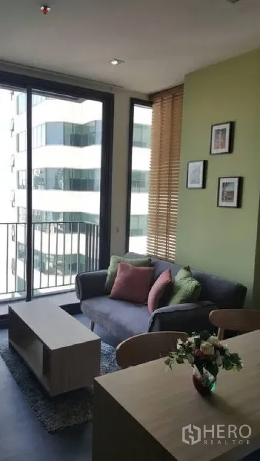 Condominium for rent in Watthana, Bangkok - Bright living room with sofa, coffee table, balcony and city view, green accent wall and wooden blinds.