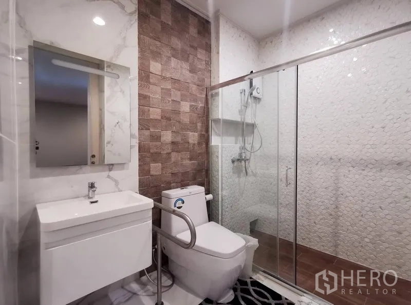 Detached House for rent or sale in Bang Kapi, Bangkok - Contemporary bathroom with glass-enclosed shower, marble tiles, textured wall, and toilet with safety rail.