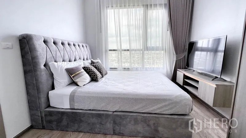 Condominium for rent in Watthana, Bangkok - Side view of master bedroom featuring upholstered bed, TV on console, full-height curtains, and skyline backdrop.