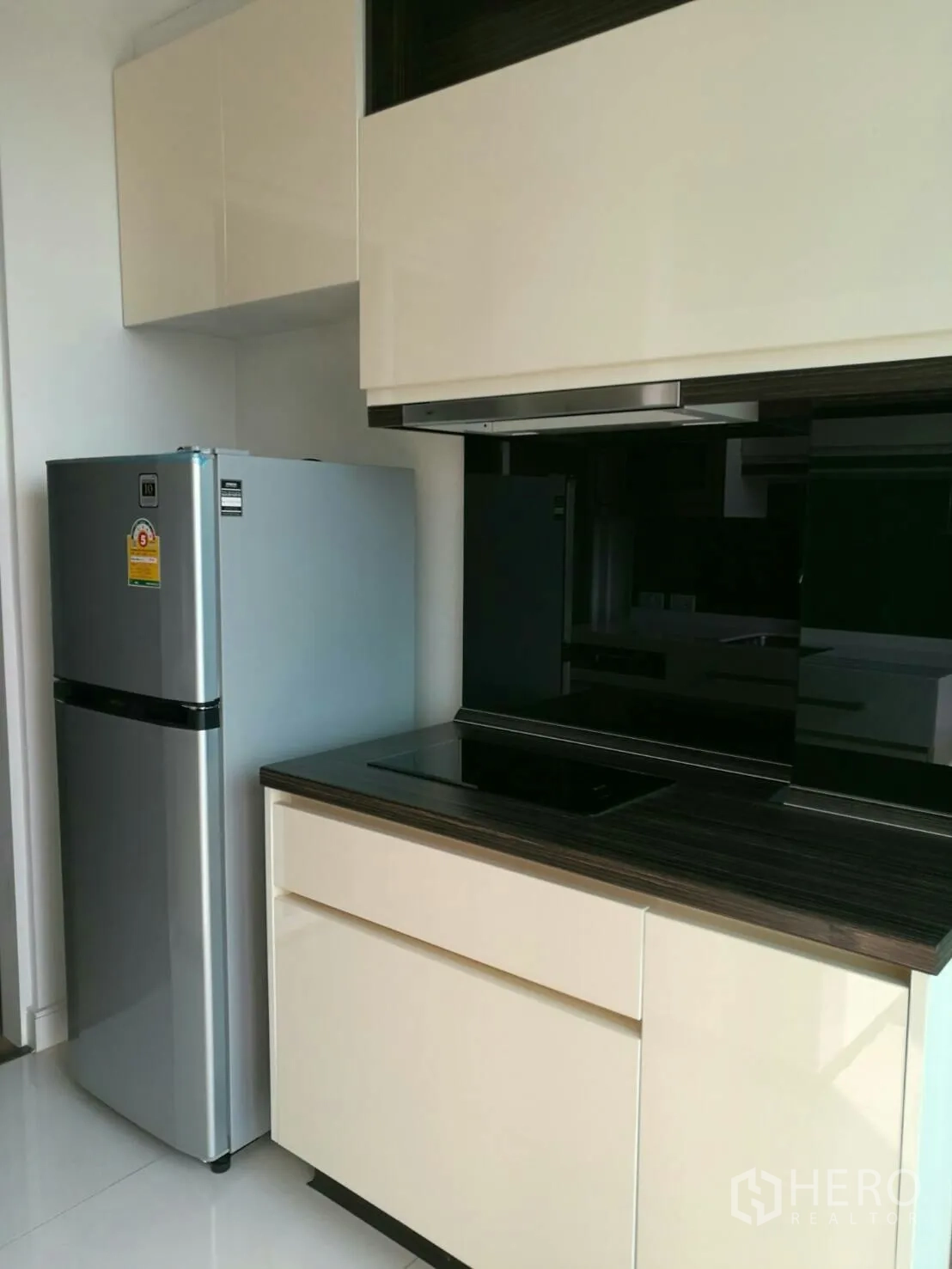 Condominium for rent in Phra Khanong, Bangkok - Compact kitchenette with silver fridge, induction cooktop, glossy cabinets and a black glass backsplash.