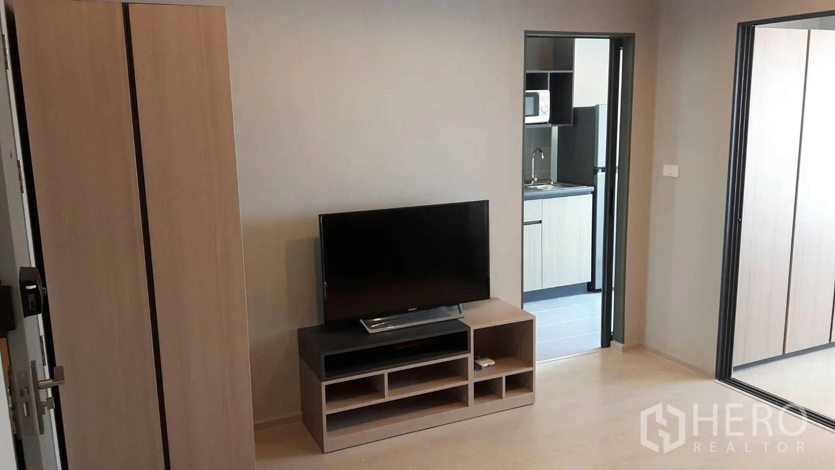 Condominium for rent in Samut Prakan - Open-plan lounge with TV console and wardrobe facing a separate kitchen in a 1-bedroom condominium.