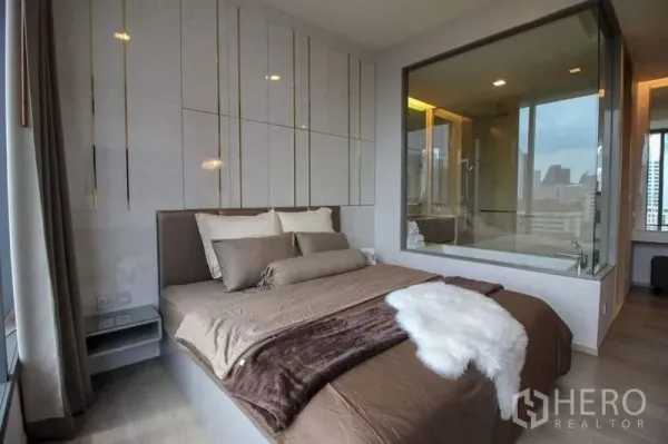 Condominium for sale in Watthana, Bangkok - Primary bedroom with upholstered bed, floor-to-ceiling window and a glass wall looking into the ensuite bathroom with bathtub and city skyline beyond.