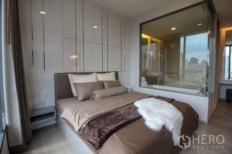 Condominium for sale in Watthana, Bangkok - Primary bedroom with upholstered bed, floor-to-ceiling window and a glass wall looking into the ensuite bathroom with bathtub and city skyline beyond.