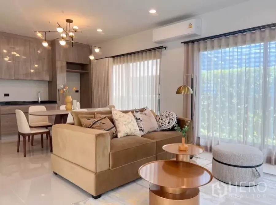 Detached House for rent or sale in Bang Kapi, Bangkok - Bright open-plan living room with beige sofa, gold coffee tables, and floor-to-ceiling curtains beside a modern kitchen.