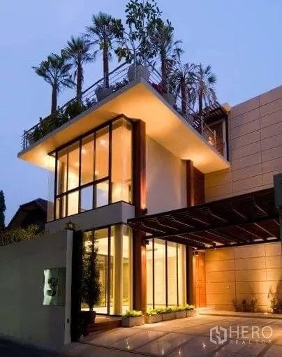 Detached House for rent in Khlong Toei, Bangkok - Modern three‑storey house exterior with floor‑to‑ceiling windows and rooftop greenery at dusk.