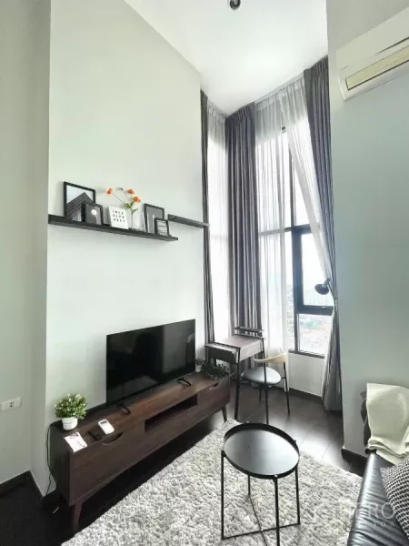 Condominium for rent in Watthana, Bangkok - TV console and wall shelf beside tall curtains and a compact work desk by the window.