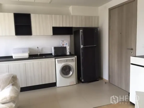 Condominium for rent in Watthana, Bangkok - Linear kitchen with light wood cabinets, black countertop, fridge, microwave, and washing machine.