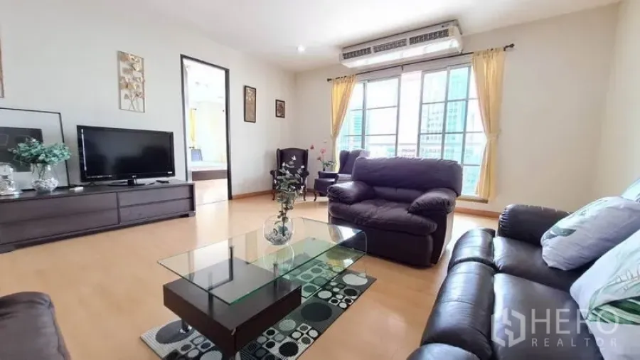 Condominium for rent in Khlong Toei, Bangkok - Bright living room with leather sofas, glass coffee table, TV console, wood floors, and large windows with city skyline views.
