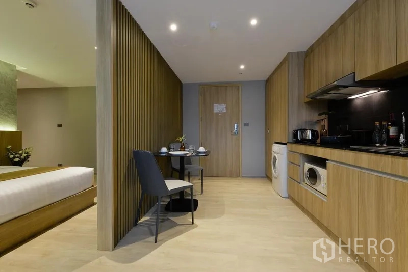 Apartment for rent in Khlong Toei, Bangkok - Open-plan layout showing kitchenette with black backsplash, washing machine and dining table by wood slats.