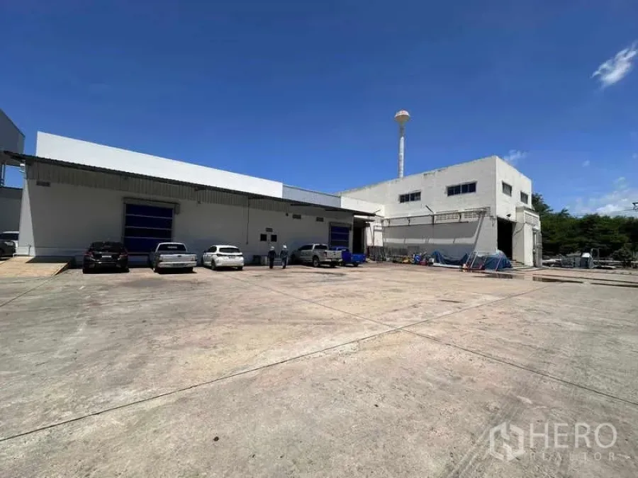 工厂 出租 - 工厂外观，蓝色卷门，大型混凝土堆场与停车区，晴空万里。