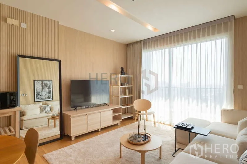 Condominium for sale in Khlong Toei, Bangkok - Bright living room with floor-to-ceiling sheer curtains, TV console, and light-wood furniture opening to a balcony view.