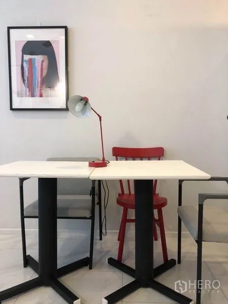 Condominium for sale in Watthana, Bangkok - Minimalist dining or work nook with a white table, red chair, desk lamp and framed artwork on a white wall.