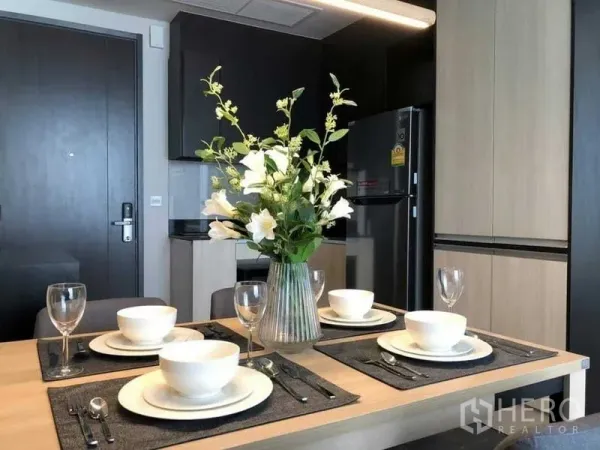 Condominium for rent in Watthana, Bangkok - Dining table set with white dinnerware and a vase, next to a sleek kitchenette with fridge and cabinets.