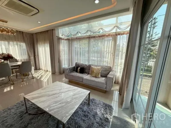 独立式住宅 出售在Bang Khen, Bangkok - 采光充足的客厅，转角落地窗，灰色沙发与大理石茶几配地毯。