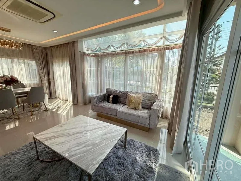 独立式住宅 出售在Bang Khen, Bangkok - 采光充足的客厅，转角落地窗，灰色沙发与大理石茶几配地毯。