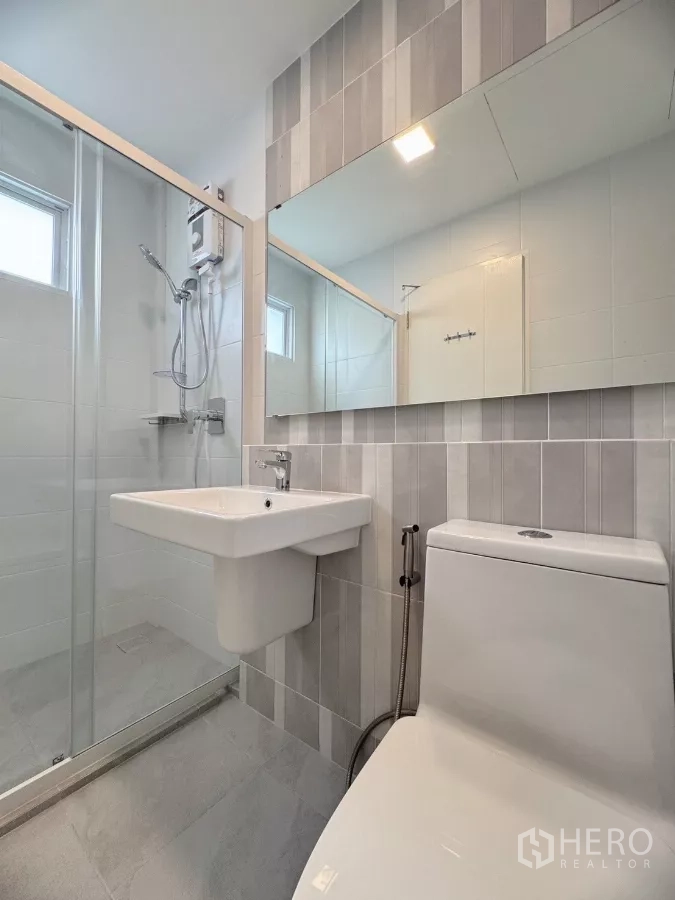 联排别墅 出租在Prawet, Bangkok - 浴室配玻璃淋浴间、灰色墙砖与镜子。