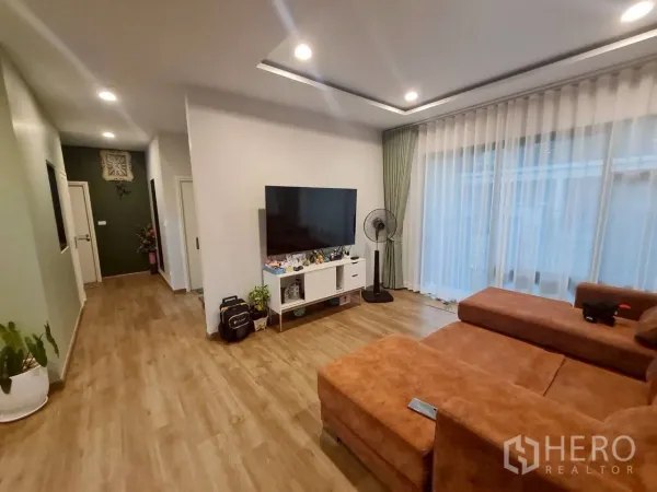 บ้าน ให้เช่าหรือขายในLam Luk Ka, Pathum Thani - มุมนั่งเล่นครอบครัว โซฟา L‑shape ตู้วางทีวี และผ้าม่านโปร่งเต็มผนัง