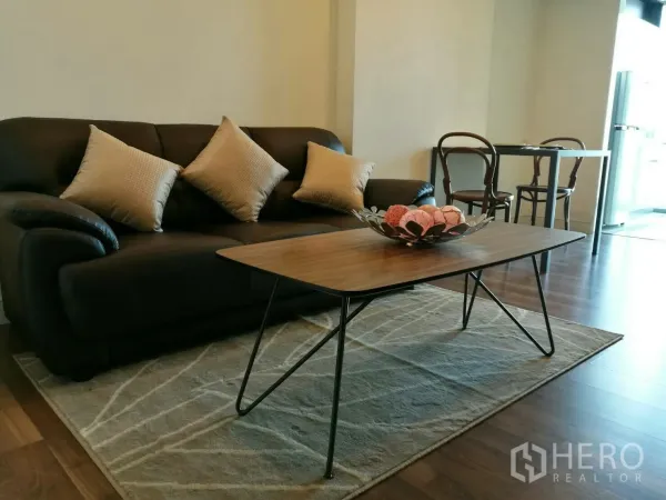 Condominium for rent in Phra Khanong, Bangkok - Modern living room with dark leather sofa, wooden coffee table and a small dining set on wood flooring in a Bangkok condo.