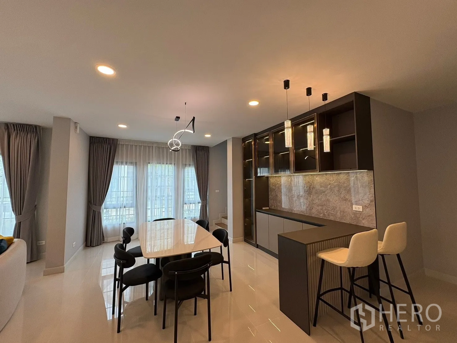 Detached House for rent in Bang Phli, Samut Prakan - Dining area with six-seat table, pendant lights, breakfast bar and glass-front cabinetry beside tall windows.