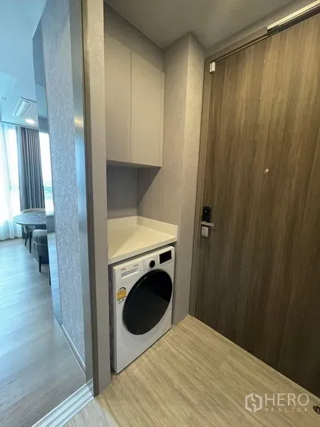 Condominium for rent in Samut Prakan - Entry nook with built-in cabinet and a front-load washing machine beside the wooden door.